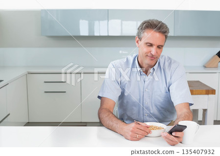 Mature male holding smartphone while eating cereal from bowl with coffee mug at kitchen countertop Mature male holding smartphone while eating cereal from bowl with coffee mug at kitchen countertop 135407932