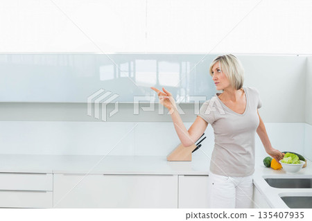 Woman standing and pointing left on countertop in white kitchen with knife block, copy space Woman standing and pointing left on countertop in white kitchen with knife block, copy space 135407935