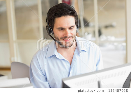 Man wearing light blue shirt and headset speaking into microphone using two monitors at office desk Man wearing light blue shirt and headset speaking into microphone using two monitors at office desk 135407938