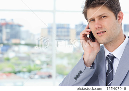 Male business professional talking on smartphone while standing by large office window, copy space Male business professional talking on smartphone while standing by large office window, copy space 135407946