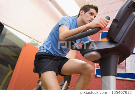 Man pedaling on exercise bike with digital console at gym wearing sky-blue shirt and black shorts Man pedaling on exercise bike with digital console at gym wearing sky-blue shirt and black shorts 135407953