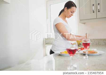 Woman slicing fresh tomatoes and arranging salad bowl on countertop in kitchen with wine glasses Woman slicing fresh tomatoes and arranging salad bowl on countertop in kitchen with wine glasses 135407954