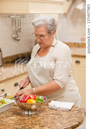 Senior woman chopping colorful peppers on cutting board on granite kitchen island with colander 135407963