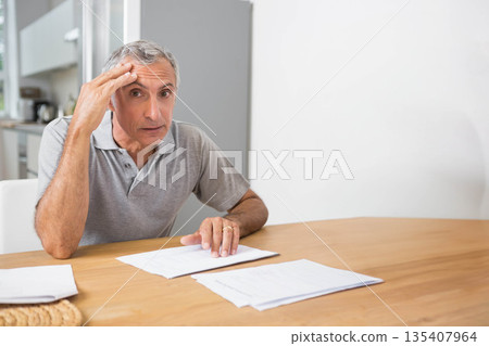 Mature male sitting at dining table reviewing paperwork with kitchen appliances visible, copy space 135407964