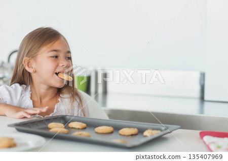 Hispanic girl eating cookie while sitting at white kitchen counter with baking tray, copy space Hispanic girl eating cookie while sitting at white kitchen counter with baking tray, copy space 135407969