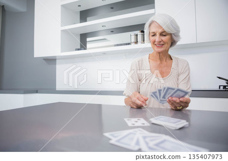 Senior woman holding playing cards while sitting at gray kitchen island with deck, copy space 135407973