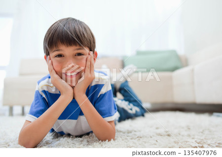 Boy lying on plush white carpet in living room, propping chin on hands near light-colored sofa Boy lying on plush white carpet in living room, propping chin on hands near light-colored sofa 135407976