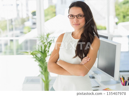 Professional woman standing with arms crossed by desk in office with monitor keyboard and plant Professional woman standing with arms crossed by desk in office with monitor keyboard and plant 135407978