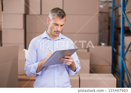 Man wearing light blue shirt and trousers inspecting boxes with clipboard and pen in warehouse 135407983