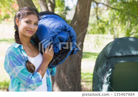 Woman carrying rolled sleeping bag over shoulder in grassy park clearing near green camping tent Woman carrying rolled sleeping bag over shoulder in grassy park clearing near green camping tent 135407984