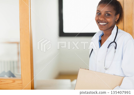 African American woman doctor with stethoscope standing by window holding cardboard box, copy space 135407991