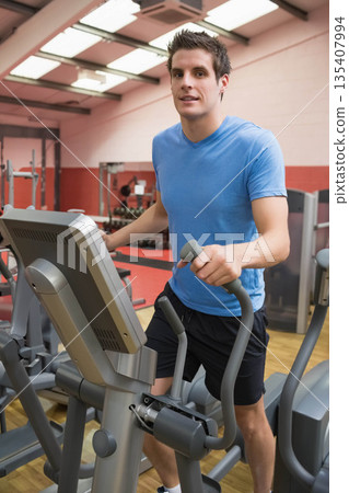 Man exercising on elliptical machine with digital display and handlebars in gym with dumbbell rack 135407994