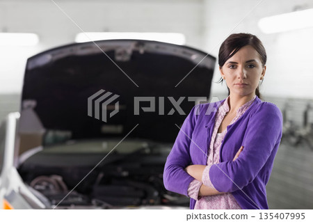 Woman late twenties crossing arms in garage near car with open hood and engine, copy space 135407995