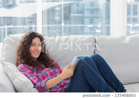 Woman in late twenties reclining on light-colored sofa in apartment living room holding tablet 135408003
