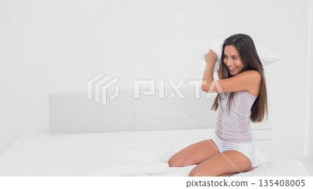 Woman kneeling on bed holding pillow overhead while wearing sleepwear in bright bedroom, copy space 135408005
