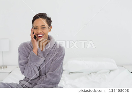 African American woman sitting on white bed wearing grey bathrobe calling on smartphone, copy space African American woman sitting on white bed wearing grey bathrobe calling on smartphone, copy space 135408008