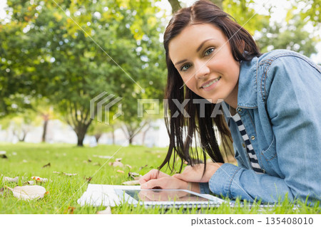 Woman wearing denim jacket reclining on park lawn, using tablet, notebook and pen copy space 135408010