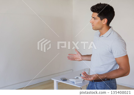 Man gesturing beside white table with telephone remote keyboard in empty room, copy space Man gesturing beside white table with telephone remote keyboard in empty room, copy space 135408021