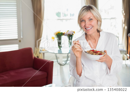 Woman smiling while holding bowl of cereal with raspberries and spoon in dining area by window 135408022