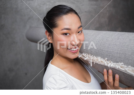 Korean woman carrying rolled-up grey rug over shoulder against smooth grey concrete wall indoors Korean woman carrying rolled-up grey rug over shoulder against smooth grey concrete wall indoors 135408023