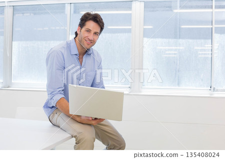 Man working on silver laptop while sitting at white desk in modern office wearing business attire 135408024