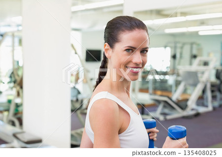 Woman gripping blue dumbbells and smiling while standing in fitness center with weight machines 135408025