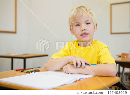 School-age boy sitting at classroom desk holding blue pencil above notebook with colored pencils School-age boy sitting at classroom desk holding blue pencil above notebook with colored pencils 135408029