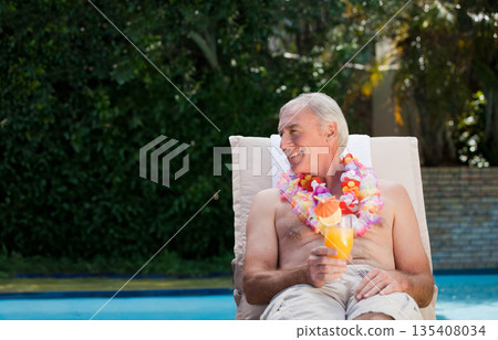 Senior man reclining on lounge chair beside pool wearing lei and holding cocktail glass, copy space Senior man reclining on lounge chair beside pool wearing lei and holding cocktail glass, copy space 135408034