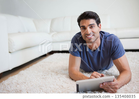 Middle-aged man lying on fluffy white carpet in living room holding tablet computer Middle-aged man lying on fluffy white carpet in living room holding tablet computer 135408035