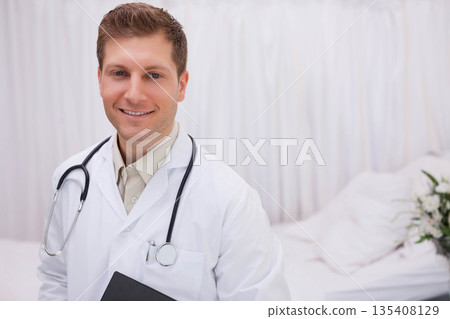 White male doctor holding folder, wearing stethoscope next to hospital bed with pillows, flowers 135408129