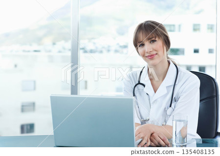 Female doctor sitting at glass desk by window using laptop with stethoscope and water, copy space Female doctor sitting at glass desk by window using laptop with stethoscope and water, copy space 135408135