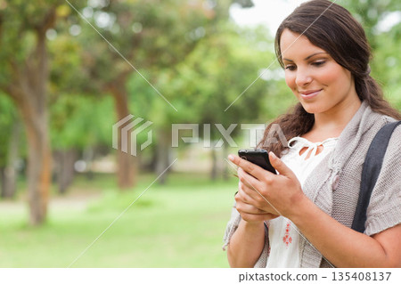 Woman holding smartphone while looking at screen in grassy park with backpack strap, copy space 135408137