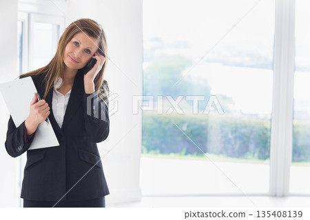 Businesswoman holding clipboard and pen and speaking on smartphone in office by windows, copy space Businesswoman holding clipboard and pen and speaking on smartphone in office by windows, copy space 135408139