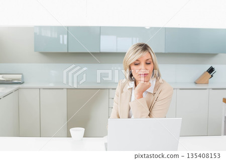 Woman working on laptop at modern home kitchen table with white mug and wooden knife block 135408153