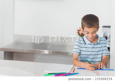 Boy drawing on white paper at kitchen counter with scattered markers and blue ruler, copy space 135408154