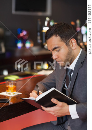 Middle Eastern man in gray suit sitting at wooden bar counter writing in notebook with drink 135408155