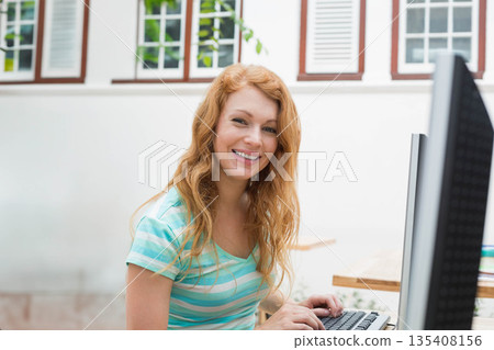 Red-haired woman sitting at home office desk typing on keyboard near shuttered window with books Red-haired woman sitting at home office desk typing on keyboard near shuttered window with books 135408156