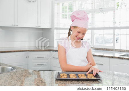 Girl wearing pink chef's hat and apron standing in kitchen touching baking tray of cookies 135408160