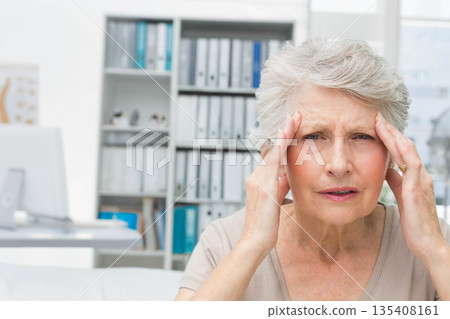 Senior female rubbing temples at desk with computer monitor and binder-filled shelves, copy space 135408161