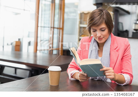 Woman wearing blazer sitting at cafe table reading paperback book with coffee cup, copy space Woman wearing blazer sitting at cafe table reading paperback book with coffee cup, copy space 135408167