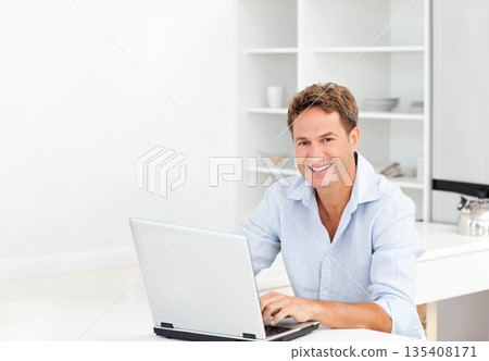 Man sitting at kitchen table typing on silver laptop wearing blue shirt with kettle, copy space 135408171