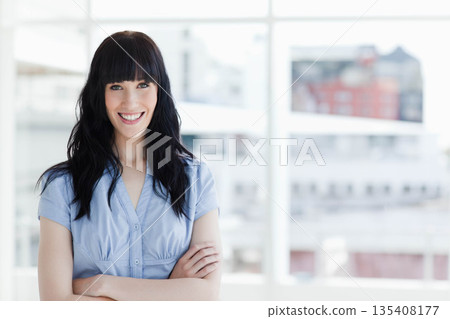 Woman standing in modern office wearing blue blouse, smiling by large windows with blurred skyline Woman standing in modern office wearing blue blouse, smiling by large windows with blurred skyline 135408177