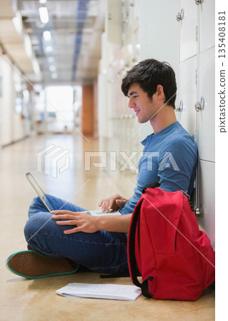Male student sitting in hallway studying on laptop next to red backpack and papers, copy space 135408181