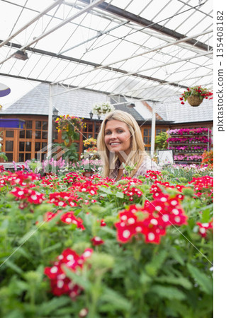 Woman standing among potted flowering plants inside greenhouse at garden center wearing plaid shirt 135408182