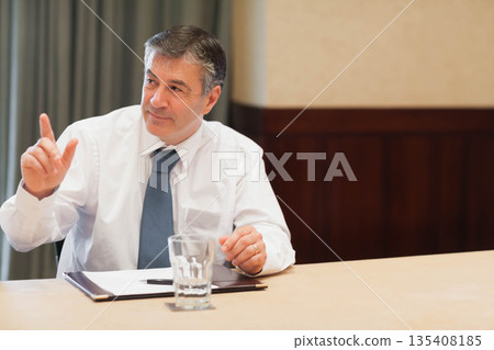 Middle-aged man wearing suit raising finger in conference room at table with folder pen water glass 135408185