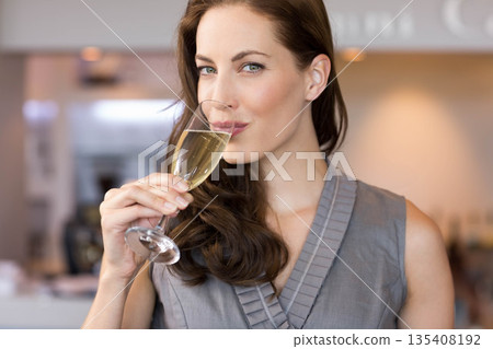 Mid adult woman in gray top holding champagne flute and sipping at bar counter with bottles 135408192