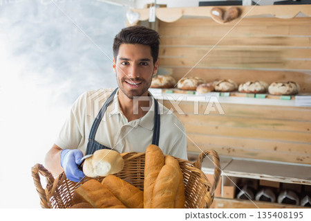 Male baker holding baguette basket wearing apron and blue glove in bakery by shelves with loaves Male baker holding baguette basket wearing apron and blue glove in bakery by shelves with loaves 135408195