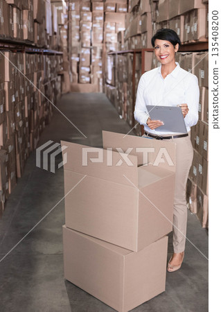Female inventory manager standing in warehouse aisle checking boxes with clipboard pen, copy space Female inventory manager standing in warehouse aisle checking boxes with clipboard pen, copy space 135408200