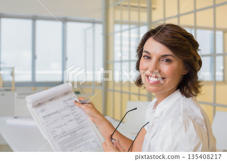 Senior woman standing at office holding clipboard documents pen and eyeglasses smiling at camera 135408217