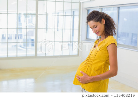 Pregnant woman standing, cradling belly in loft with grid windows plus concrete floor, copy space 135408219